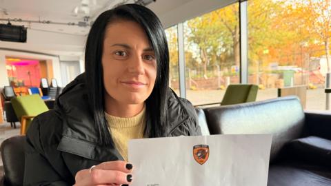 Sophie Griffiths looking at the camera holding a letter with a Hull City logo at the top. She has long black straight hair with a side parting. She is wearing a black coat and a yellow sweater. Her nails are painted black. She is sitting on a black sofa inside an office-like space.