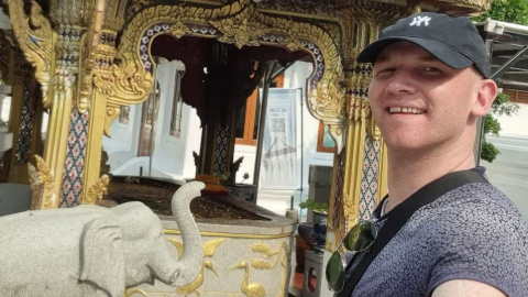 Rory McColl , who is wearing a black baseball cap, takes a selfie in front of a stone elephant and a historic building