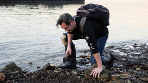 Mudlark Jason Sandy wearing a backpack is bent over and holding a trowel as he searches the shoreline of the River Thames for historical items