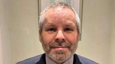 Sean McNamee. He has short silver hair and beard, wearing a dark blue blazer and check shirt. Behind him is a wall with a photo frame on it.