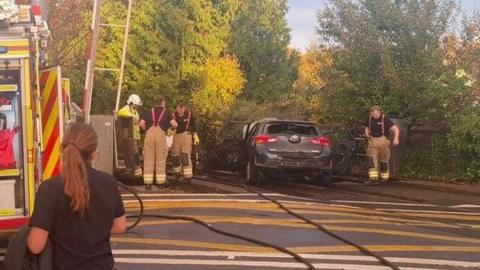 Firefighters stand near a burnt-out vehicle on a level crossing
