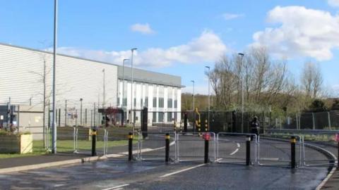 A grey industrial building behind a number of large bollards and fencing. 