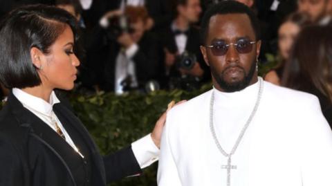 Cassie Ventura looks side on at P Diddy while attending a red carpet event. She is dressed in black and he is dressed in white