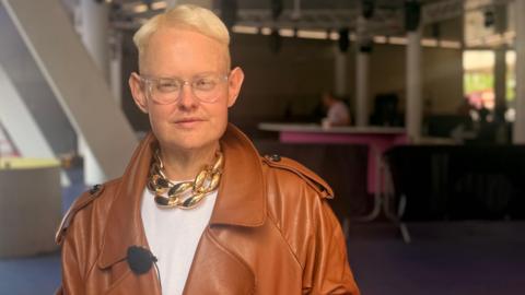 Michael Hopkinson, who is wearing a brown leather jacket, gold chain and white T-shirt, is staring directly at the camera.
