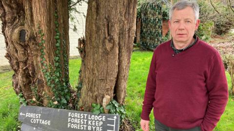 A man with short grey hair, wearing a burgundy jumper standing next to signage pointing to the holiday lets on his property