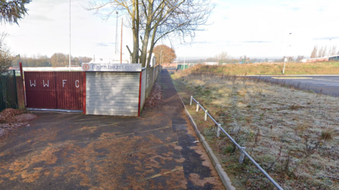 The entrance to Walsall Wood football club. It is autumn and there are leaves on the ground. There is a fence with WWFC lettering. 