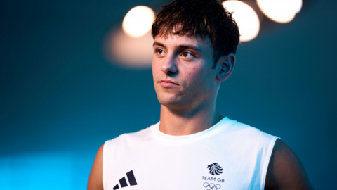 A man with brown hair stands in a Team GB white tank top with a faded blue background