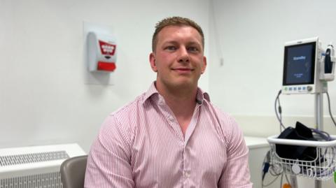 Aaron is in a striped shirt seated in a clinical room, with medical equipment and a monitor visible behind them.
