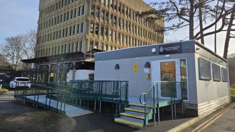 A small portacabin is outside a large office block with many windows. The sign above the door says Yeovil Police Station.