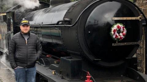 A man in a black zip-up puffer jacket stood next to a black steam train with a flower wreath attached to the front.