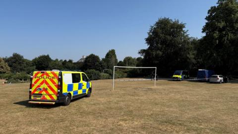 A field with a football goal in the middle. There is police tape across the field and a police van to the left. To the right is three other vehicles.
