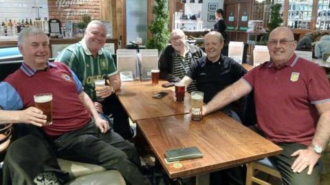 Five men sat around a wooden square table smiling and holding pint glasses of beer or larger.