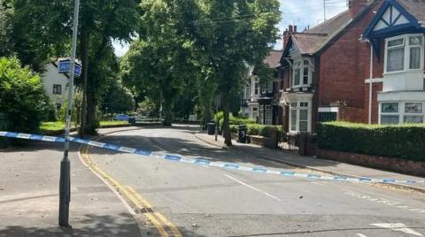 Police tape on a residential road, with trees and houses either side of the street. The tape is tied around a lamp post and stretches across the road which has double yellow lines on it. Behind it, trees line the road with red-brick houses at the side.