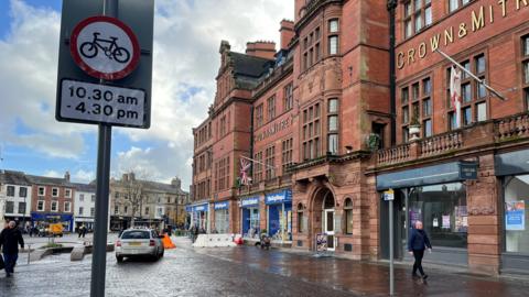 The entrance to the pedestrian area by the Crown and Mitre in Carlisle, which is a large and beautiful redbrick building with leaded windows, a first floor balcony running most of its length, ornate stone work and many chimneys. The ground floor is given over to shop fronts. A taxi is parked in front of an orange bollard, behind a sign banning cyclists from 10.30am to 4.30pm.
