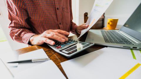 An elderly man using a calculator and laptop as he examines a paper spreadsheet, with a notebook, pen and coffee mug beside him.