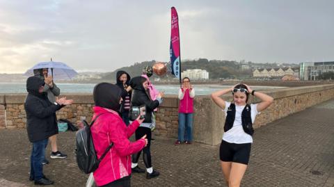 Emily is wearing a white shirt and black shorts with a hydration vest, she is in the foreground, hands on head. Several people in bright pink jackets and dark clothing stand nearby, some clapping and others taking photos. A pink event flag and a metallic balloon are visible, suggesting a celebratory moment. The background shows a stone seawall, water, and distant buildings under a gray, rainy sky.