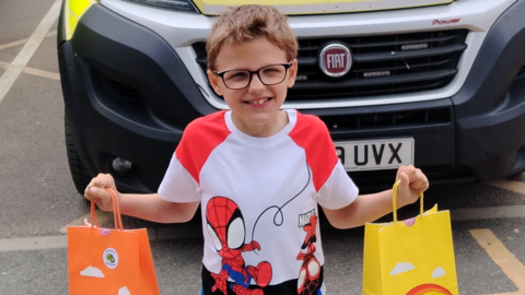 A boy in glasses and a white spider man tshirt holding two bags, one orange and the other yellow in front of an ambulance