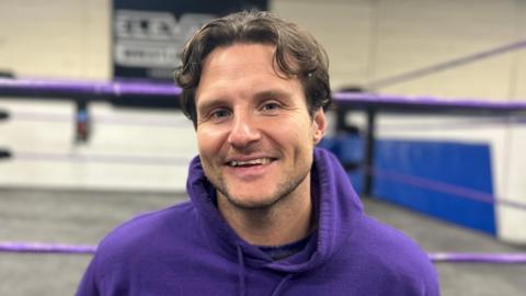 Joseph Conners smiles at the camera wearing a purple hoodie. He has brown hair and is in front of a wrestling ring