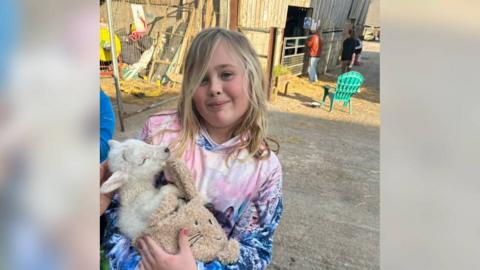 A girl with long blond hair wearing a blue and pink top is holding a lamb and a cuddly toy. She is standing in a farm yard and farm buildings are behind her as well as a blue chair.