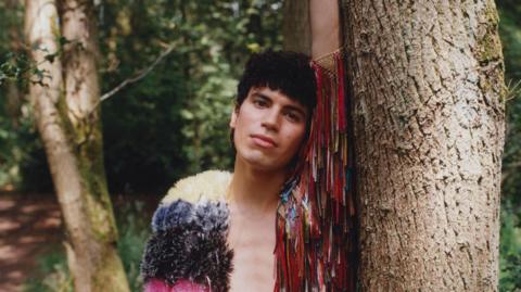 Jacob Alon leans against a tree in a lush green woodscape. They are wearing a multi-coloured jacket with tassles
