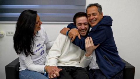 Omer Shem Tov hugging a family member and holding the hand of a woman sat on a black sofa.