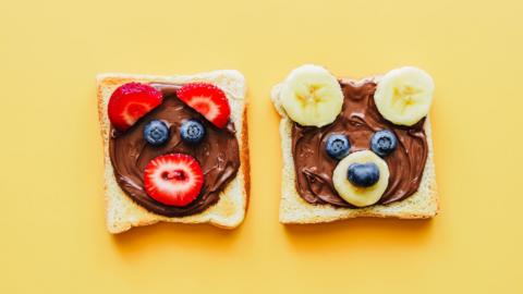 Two slices of toast with chocolate spread and fruit which is shaped to look like bears. The one on the left has strawberries for ears and mouth and blueberries for eyes and the one on the right has bananas for ears and mouth and blueberries for eyes and nose. On a yellow background