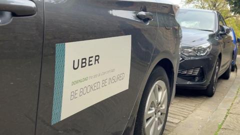An Uber car which is black and has a sign on the side saying "Uber, download the app, be booked, be insured". It is parked at the side of a path with another black car behind it.