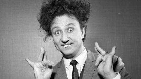 Ken Dodd, wearing a suit, with wild hair, looks at the camera, with his hands pinched in the air