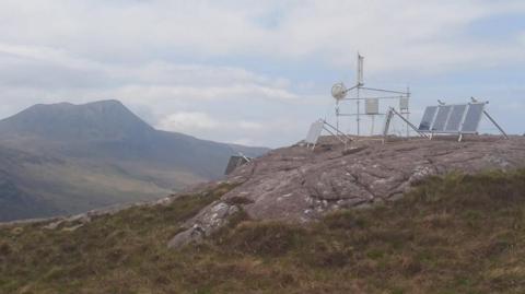 Technology equipment on a rocky hill. There are receivers and transmitters. There is grass further down and a larger mountain in the distance. A cloudy sky. 