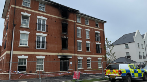 A picture of the outside of the flats. It's a large brick building, with fencing around it and a police vehicle in front it. There is signs and police tape. 