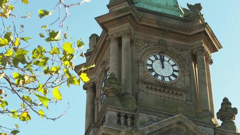 Birkenhead Town Hall clock tower with tree seen in the foreground. The black and white clock-face is showing just before midday.