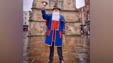 A very tall man who has a long white beard is dressed in a town crier outfit. He has a red and blue coat with gold detailing, and black trousers and is ringing a large gold bell. He is wearing a black and gold hat with a red feather.