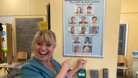 Maria smiles at the camera and points to a prop board on the set of doctors with a picture of her on it 