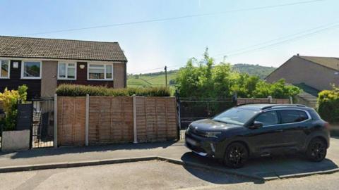 A quiet residential street with semi-detached houses featuring brick and pebble-dash exteriors, tiled roofs, and a wooden fence dividing the properties. A black SUV is parked in front of one house.
