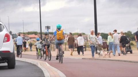 Cyclists riding on a dedicated bike path alongside a beach, separated both from the road and pedestrians, who walk on a pavement.