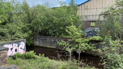 An old industrial unit with corrugated iron walls and graffiti overlooks a river with trees and greenery
