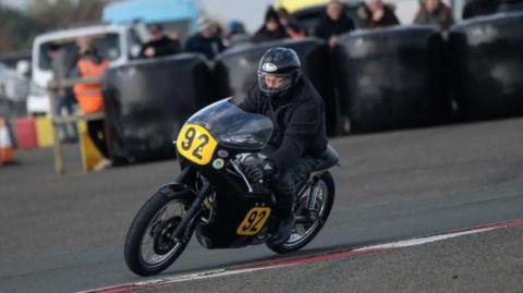 Edward Poole in black leathers and helmet riding a classic bike with yellow plates with the number 92 on them.