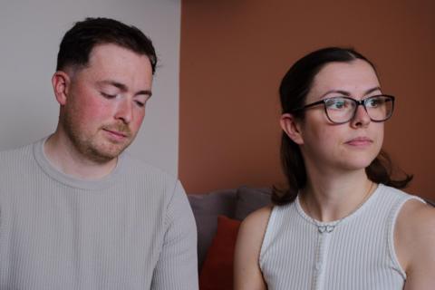The McCreadys are sitting together on a sofa. Lauryn is looking wistfully out of a window while Andrew is looking downards towards their daughter's memory box