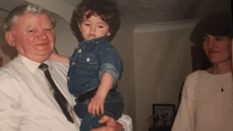 A grainy colour photo of Alec Ward holding a toddler - a young Liron Velleman. Mr Ward has grey hair and is wearing a shirt and tie. The little boy has thick curly brown hair and is wearing a denim shirt. To the right of the photo, looking on, is the boy's mother.