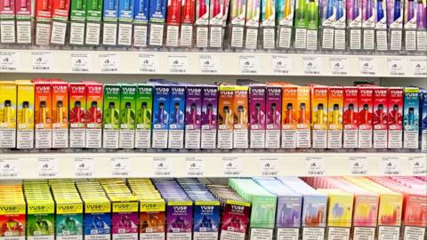 Colourful vape packaging is on display on a shelf in a shop.