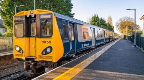 A photograph showing a blue, grey and yellow-painted Merseyrail 507 Class train at an empty station platform on a clear, bright day