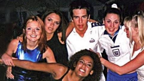 Group image of Geri Halliwell in a blue dress and Victoria Adams in a black dress on the left of Anthony in a white shirt. mel C wears a white England football top with Emma Bunton in white at the right. Mel B crowches down in front stretching out her arms.