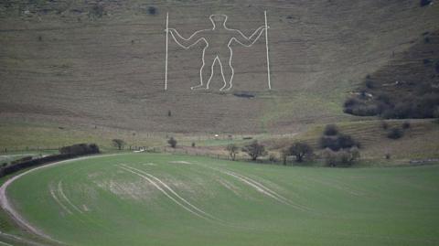 An image of the Long Man of Wilmington in Wilmington, East Sussex. The chalk man figure can be seen on the hill, carved out in white.