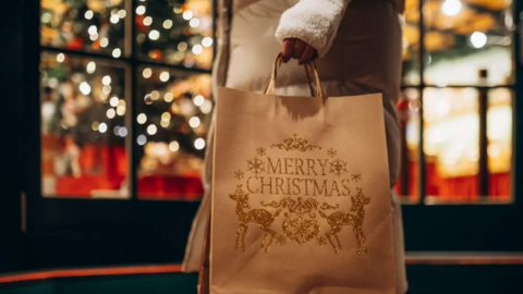 A shopper in a brown coat holds a beige bag which has Merry Christmas written on the side. They are stood outside a blurry, festive window display.