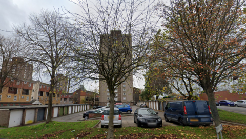 A car park with garages to the left and right.  Vehicles are parked and there is a large concrete tower block in the background