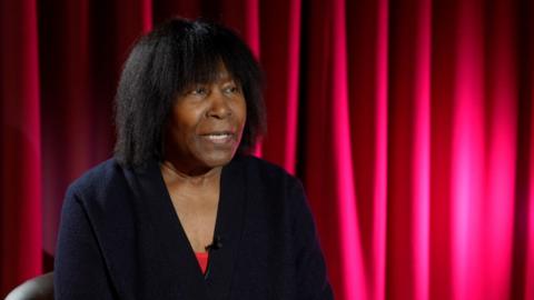 A black woman in a back jacket with black hair sat in front of a red curtain