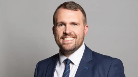 A man with brown hair and a short beard is wearing a white shirt with a navy blue suit jacket and a navy blue tie with thin yellow stripes. He is standing against a light grey backdrop.