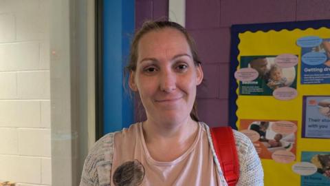 A woman with her hair tied back smiles at the camera, she is wearing a pale pink T-shirt with a cardigan, and is holding her child's rucksack. Behind the wall is painted in white, blue and purple, with a presentation board on the wall to the right.