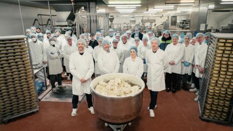 The floor of a bakery production centre, with dozens of employees standing for a group photograph alongside plenty of pastry