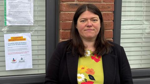 Emily Spurrell has long dark hair and is wearing a green floral patterned top and a black jacket. She is stood in front of the house window where a closure order has been attached to the window